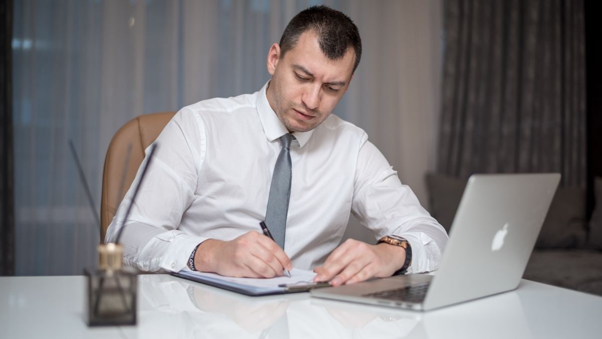 Businessman in suit writing notes at desk with laptop, reflecting modern office life.